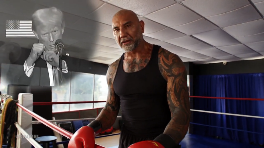 A still of Dave Bautista wearing boxing gloves at a gym while an ominous and frankly sad image of disgraced former president Donald Trump appears superimposed on the corner of the screen.